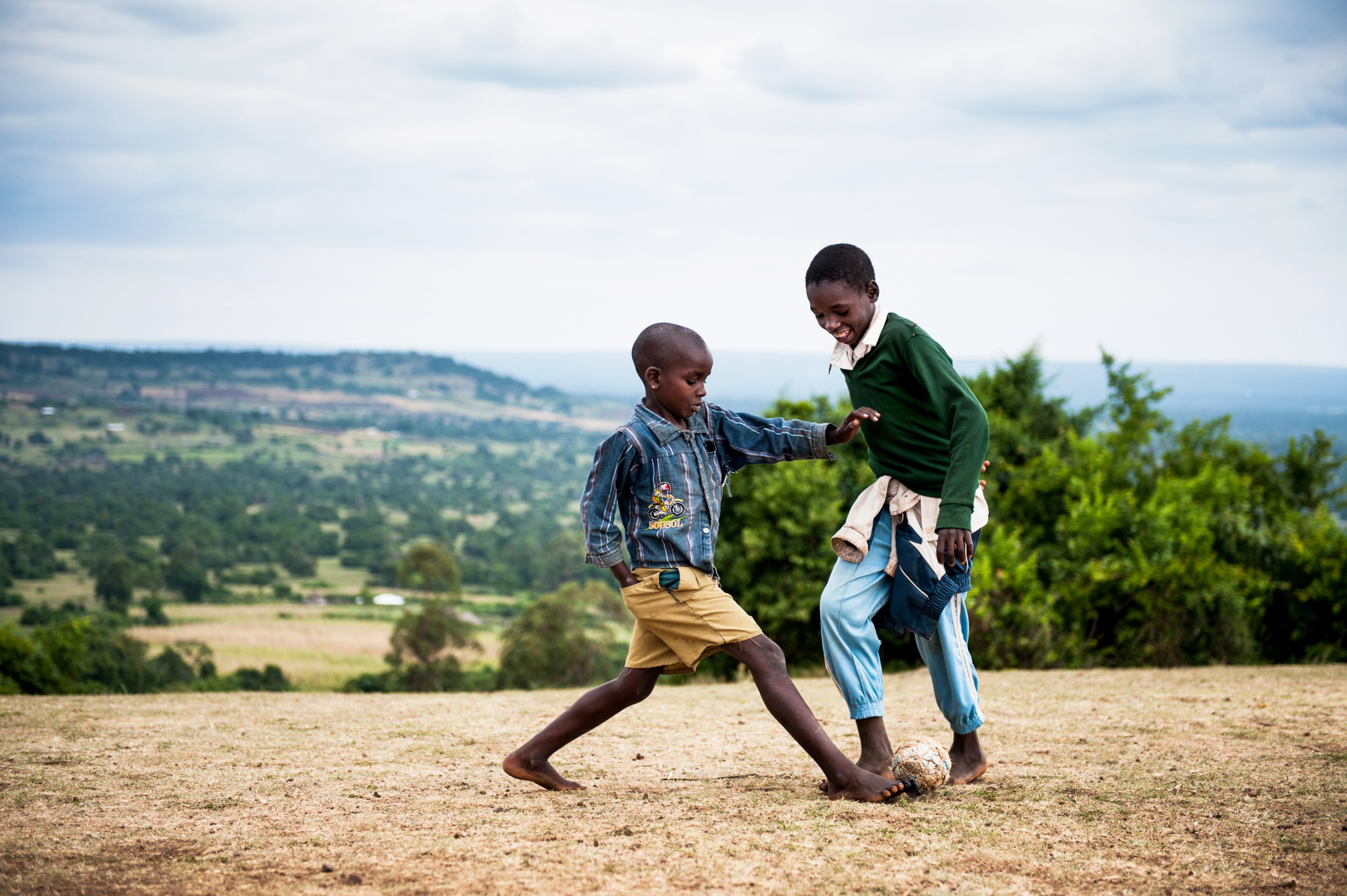 Two barefoot boys playing soccer with a worn ball on a dry grass ridge above a green valley, one lunging for the ball as the other laughs and shields it.